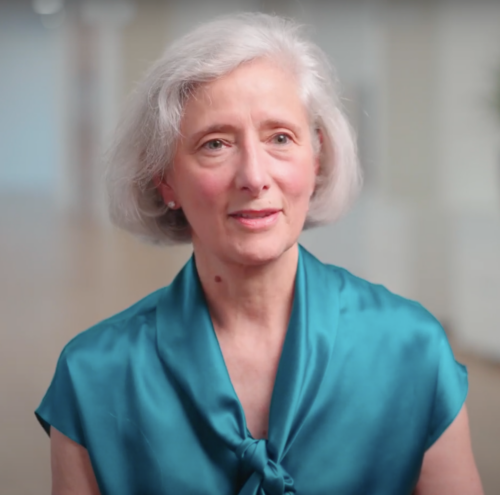 Pamela Dippel Choney, an older woman with short white hair, stands gracefully in a teal blouse against a softly blurred indoor backdrop.