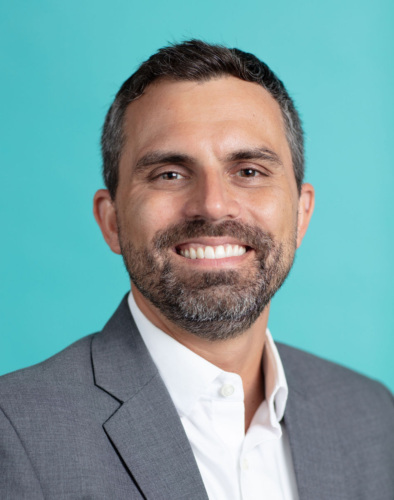 Man with short dark hair and beard wearing a gray suit jacket and white shirt, smiling in front of a plain turquoise background.