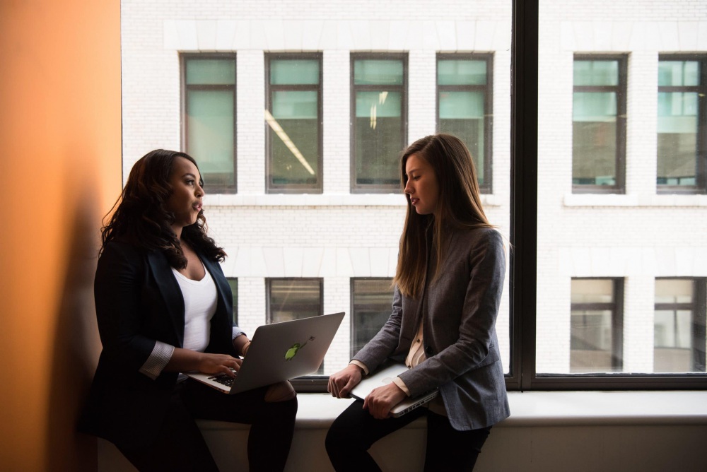 Two women discuss work with laptops by the window.