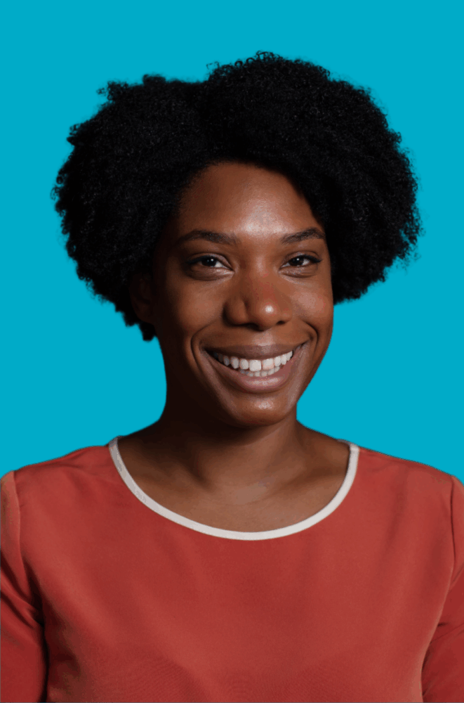 A person with short, curly black hair smiles at the camera, wearing a coral top with a white trim, against a solid turquoise background.