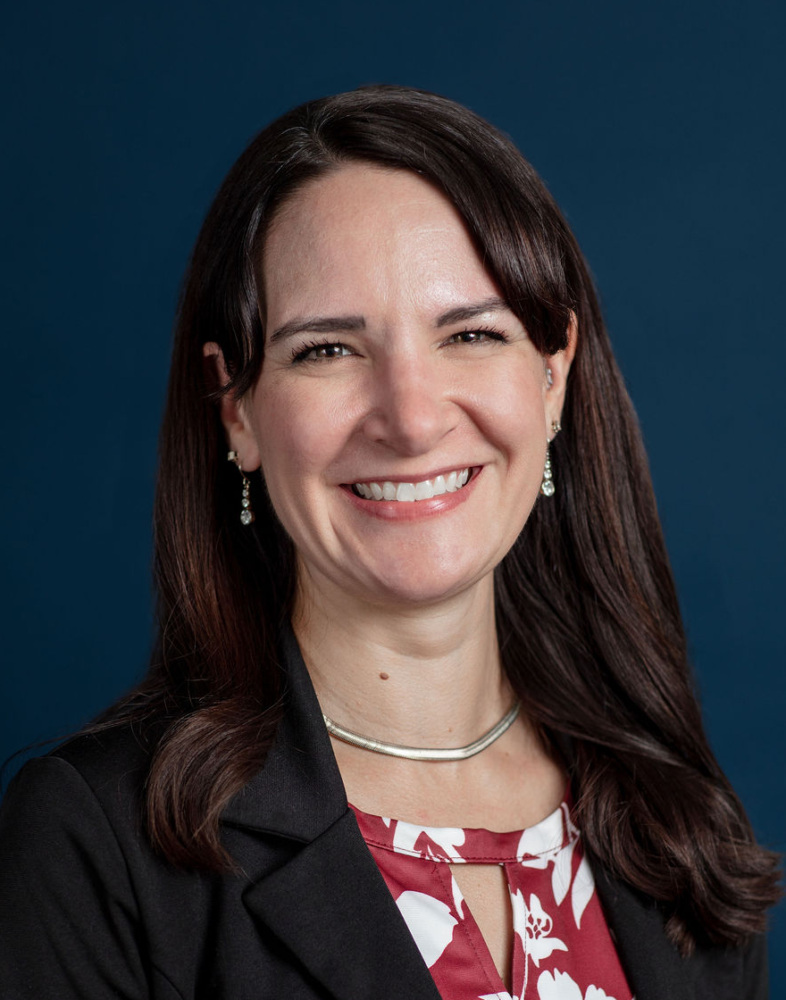 Woman with long brown hair wearing a black blazer and red and white floral blouse, smiling in front of a dark blue background.