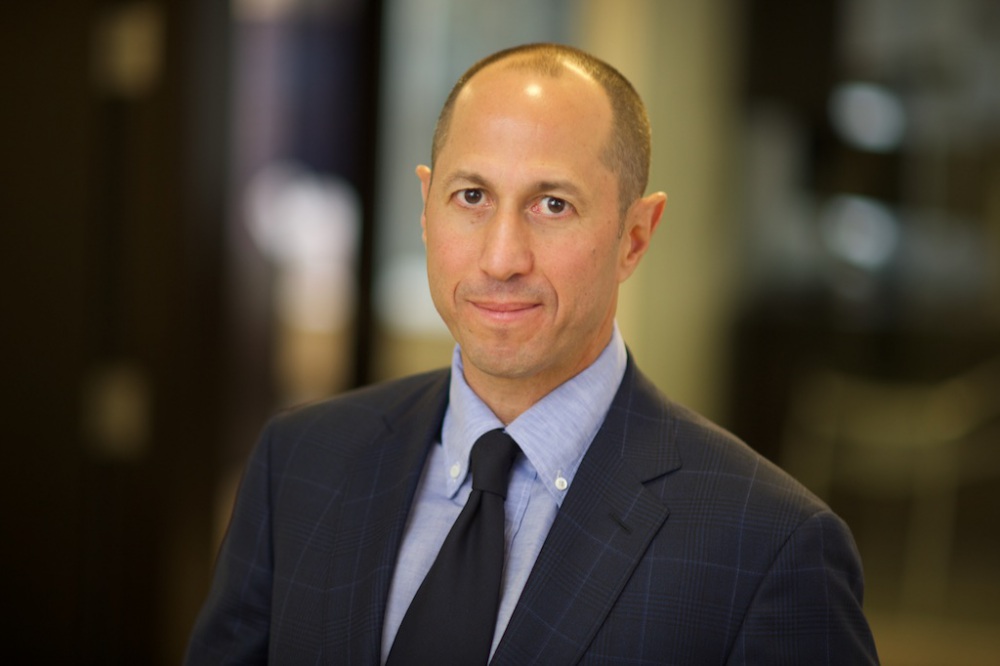 Headshot of Marc Spilker wearing a black suit with a blue dress shirt and black tie.