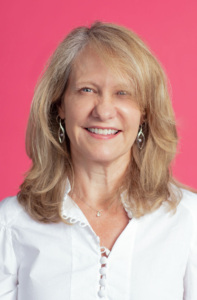A woman in a white shirt is smiling in front of a pink background.
