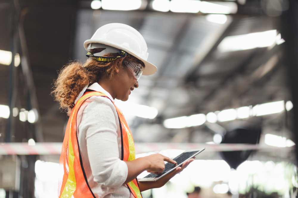 A woman worker in a hardhat stands in a distribution center viewing a tablet.