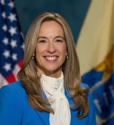 Woman with long, light brown hair wearing a blue blazer and white blouse, smiling in front of U.S. and state flags, representing the spirit of the New Jersey Pay It Forward initiative.