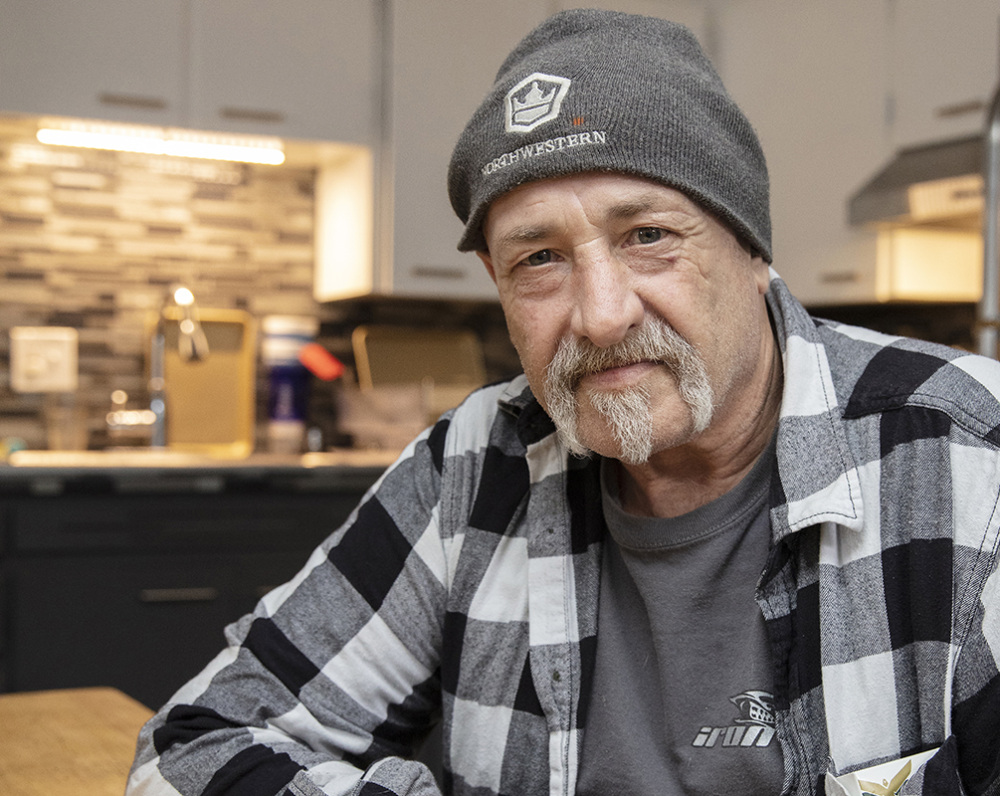 An elderly man in a gray flannel and gray beanie hat poses for the camera.