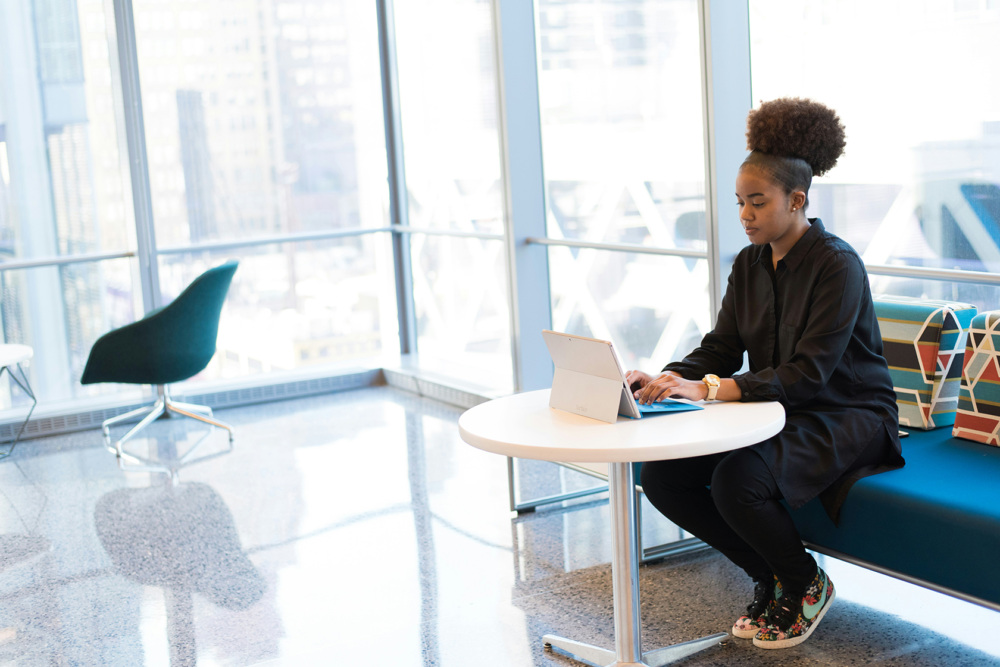 A person sits on a blue sofa in a modern office, working on a laptop at a round white table.