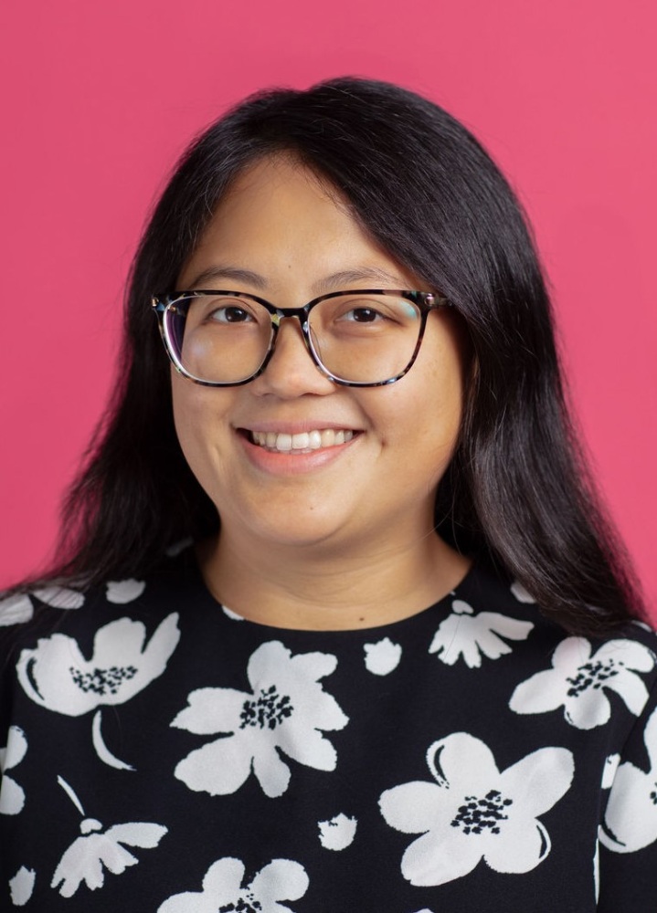 Asian woman in glasses smiling in front of a pink background.