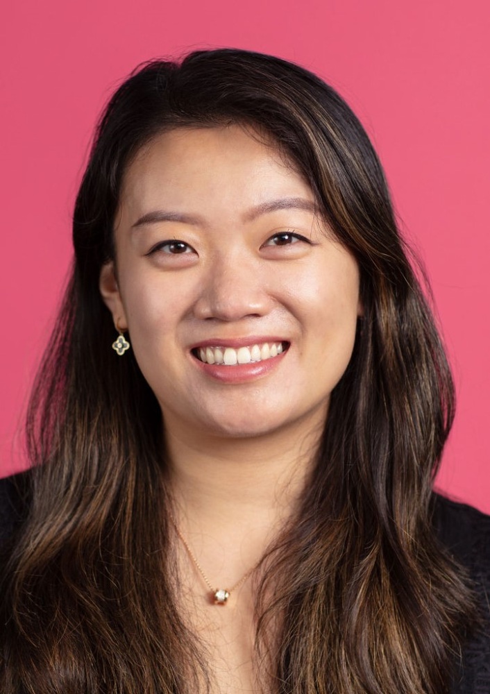 An asian woman smiling in front of a pink background.