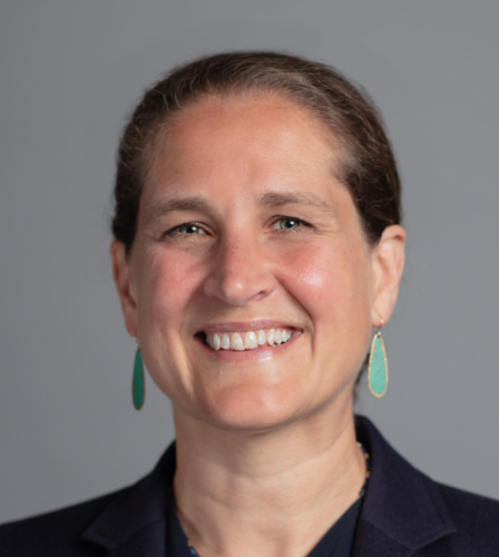 A smiling woman in a blue jacket and blue earrings.