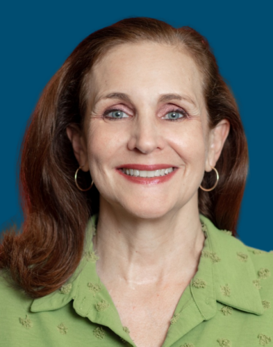 A middle-aged woman with long brown hair, wearing a green blouse and gold hoop earrings, smiles at the camera against a solid blue background.