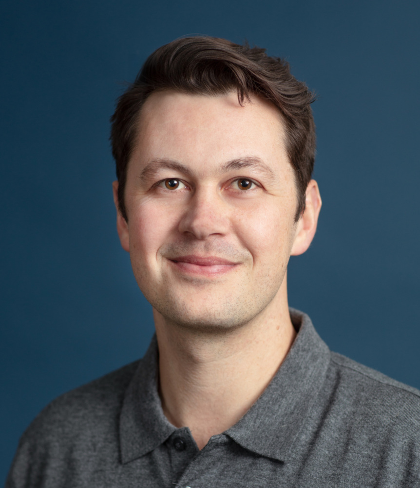 A person with short dark hair wearing a gray polo shirt against a blue background, smiling slightly.