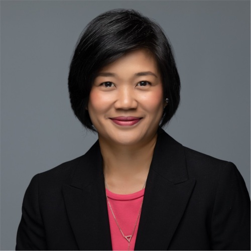 Portrait of a person with short black hair, wearing a black blazer and pink top, smiling against a gray background.