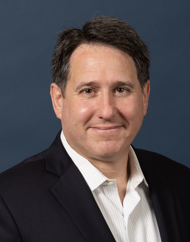 Middle-aged man with short dark hair, smiling, wearing a dark blazer and white shirt, against a blue background.