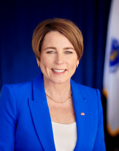 Portrait of a smiling woman with short brown hair, wearing a blue blazer and a white top, posing in front of a blue backdrop with a flag.