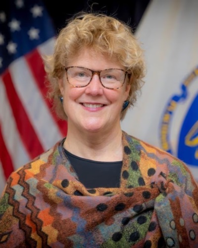 A woman with curly blond hair and glasses smiles, wearing a colorful shawl; an american flag and official seal in the background.
