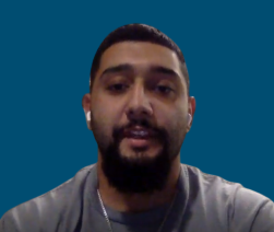 A man with a trimmed beard and mustache, wearing a gray shirt and white earphones, is positioned in front of a plain blue background.