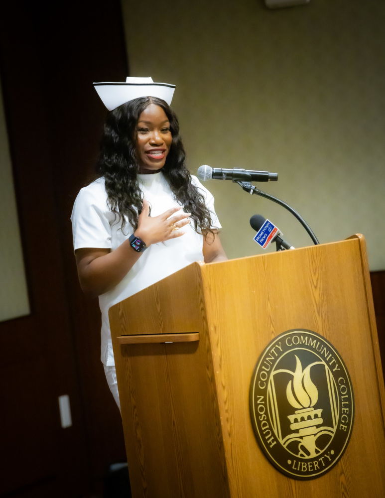 A person stands at a podium with a Hudson County Community College emblem, wearing a white nursing uniform and cap, speaking into a microphone.