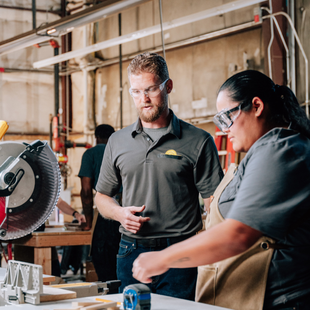Two individuals wearing safety goggles work in a workshop. One person, standing and wearing a gray polo shirt, appears to be instructing or discussing with the other person, who is standing and working.
