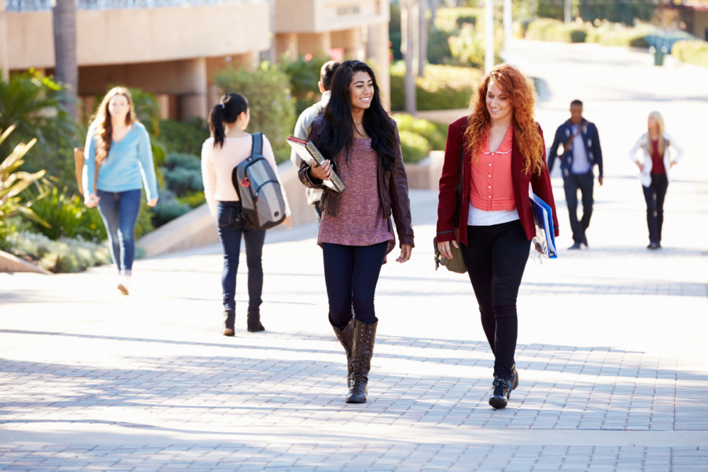 Two women walking and talking on a college campus pathway, holding books and smiling. Other students are walking and talking in the background.