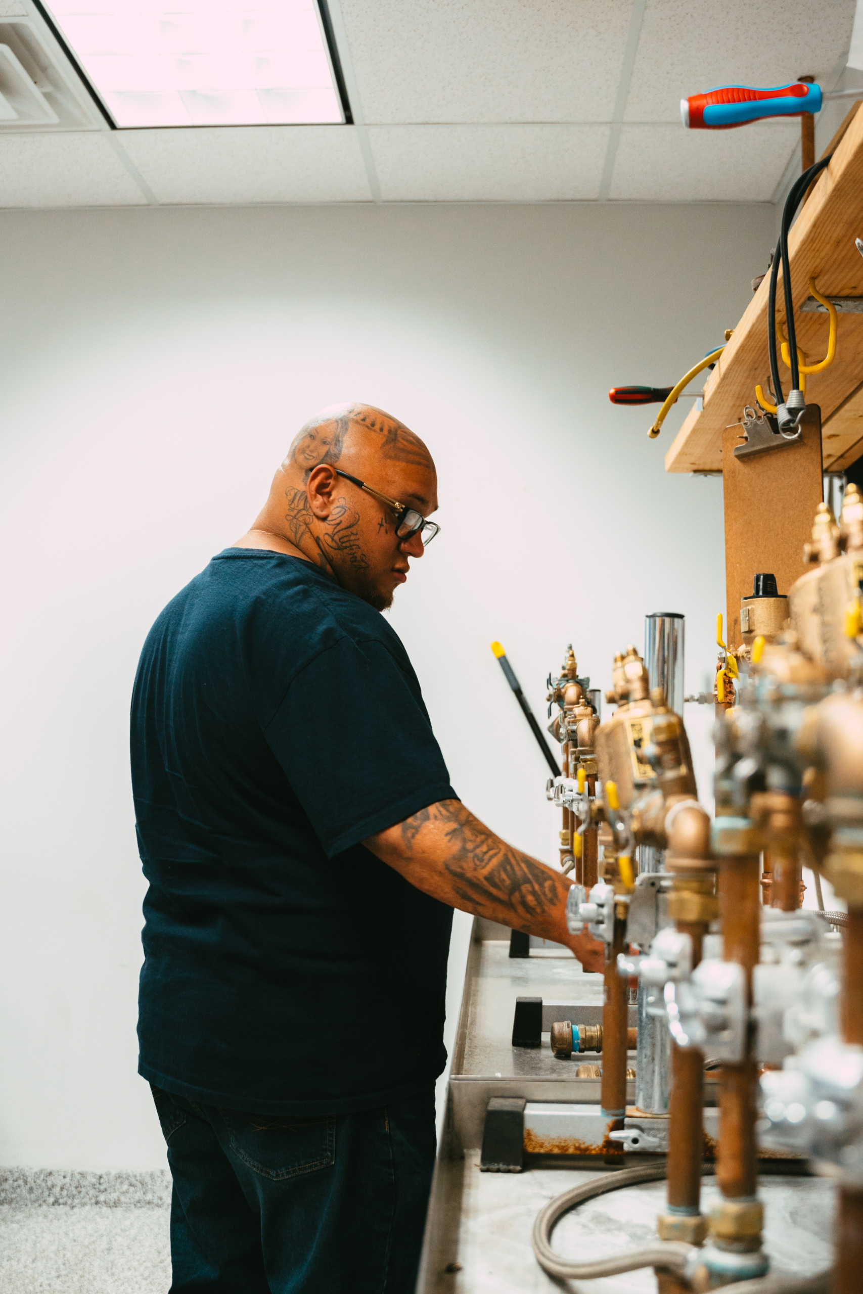 A person with tattoos operates plumbing equipment in a workshop with pipes and valves on a workbench.