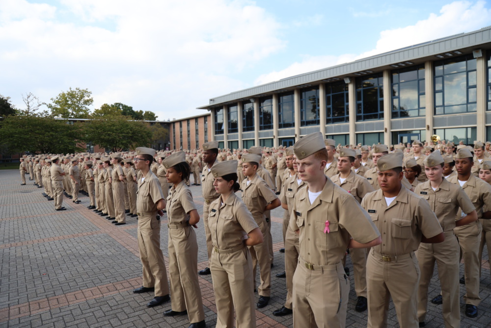 A group of people in uniform stand in formation on a paved area outside a building.