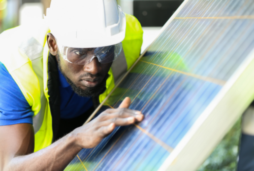 A worker wearing a hard hat, safety glasses, and a hi-vis vest inspects a solar panel closely, touching the surface with his hand.