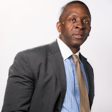 A person wearing a pinstripe suit, blue shirt, and beige tie, standing against a white background.