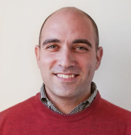 A man with a shaved head wearing a red sweater over a checkered shirt smiles in front of a plain background.