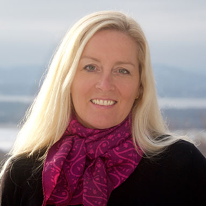 A woman with long blonde hair wearing a pink patterned scarf and a black top smiles at the camera against a blurry outdoor background.