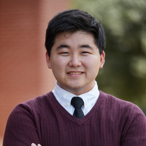 A person with short dark hair, wearing a maroon sweater, white shirt, and black tie, stands in an outdoor setting with a blurred background.