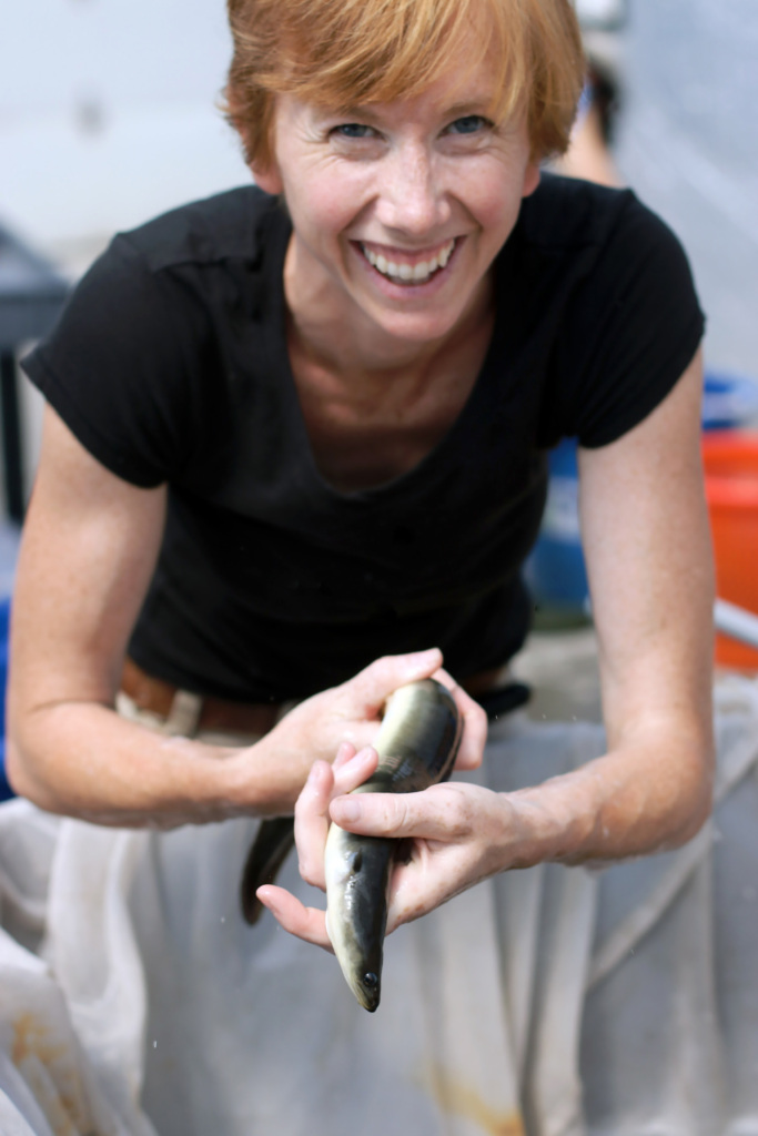 Person holding a fish with both hands, smiling, wearing a black shirt.