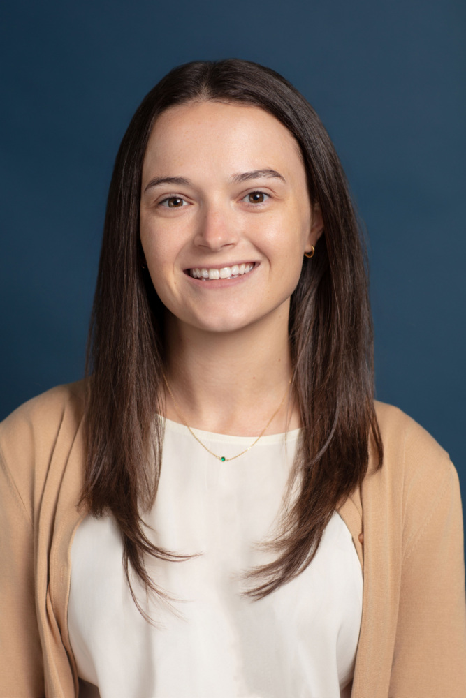 A woman with straight brown hair, wearing a beige cardigan over a white top, smiles at the camera against a plain dark blue background.