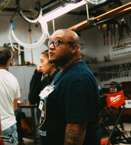 A man with tattoos and glasses stands in a workshop, surrounded by tools and equipment.