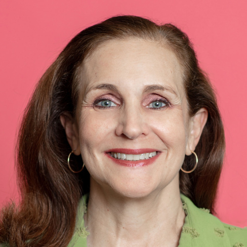 A person with long brown hair and hoop earrings smiles against a pink background.