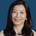 Portrait of a woman with long dark hair, wearing a sleeveless top, smiling against a plain blue background.