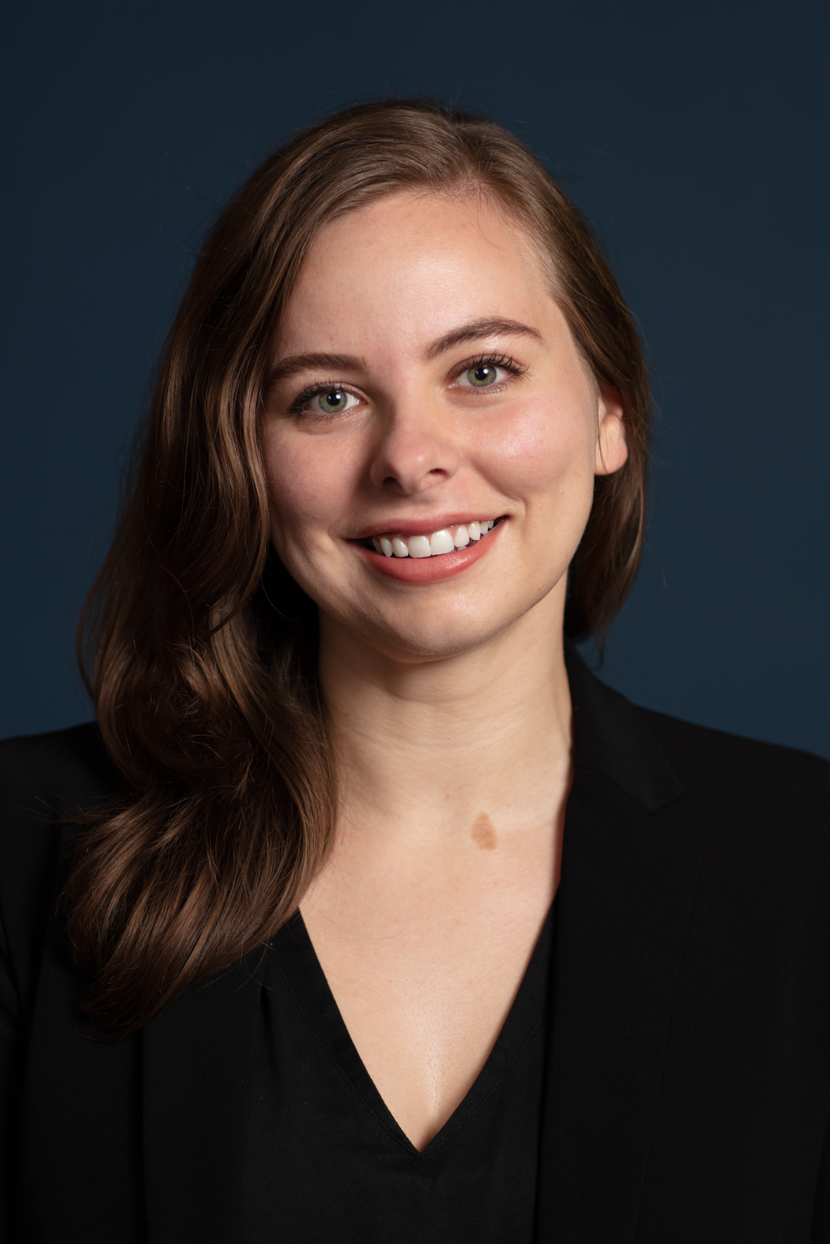 A woman with long brown hair smiles, wearing a black blazer against a dark blue background.