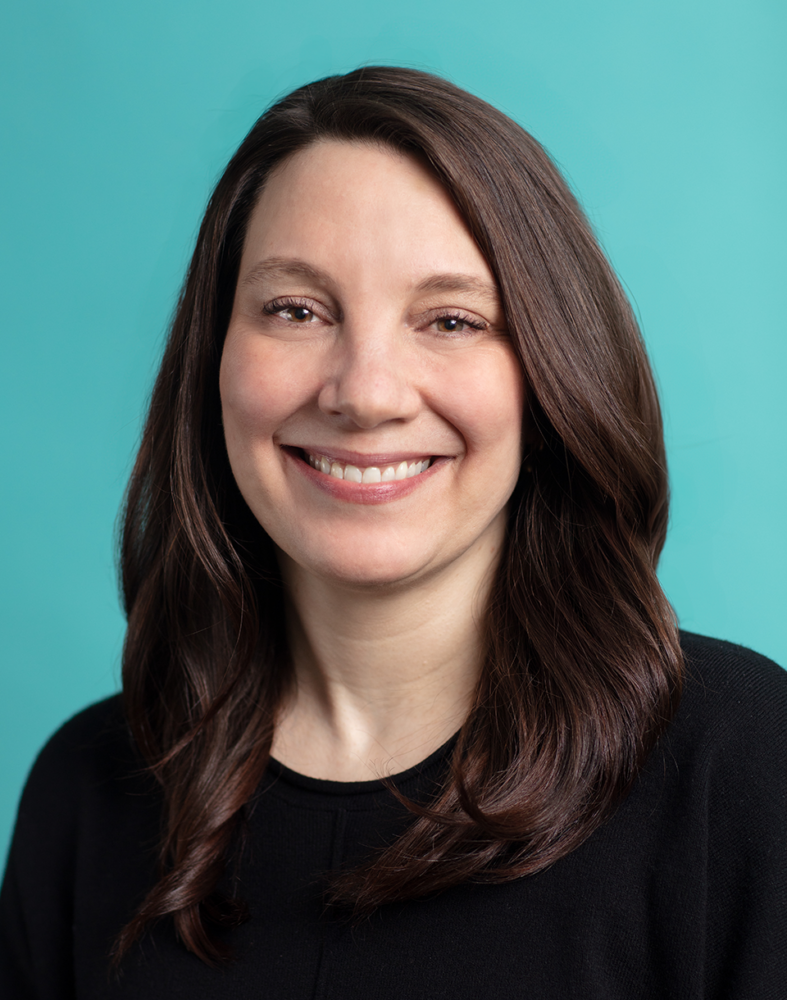 A person with long brown hair and a black shirt smiles against a turquoise background.
