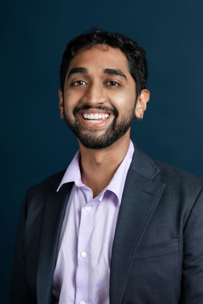 A man with short dark hair and a beard, wearing a gray suit jacket and a light purple shirt, smiles at the camera against a dark blue background.