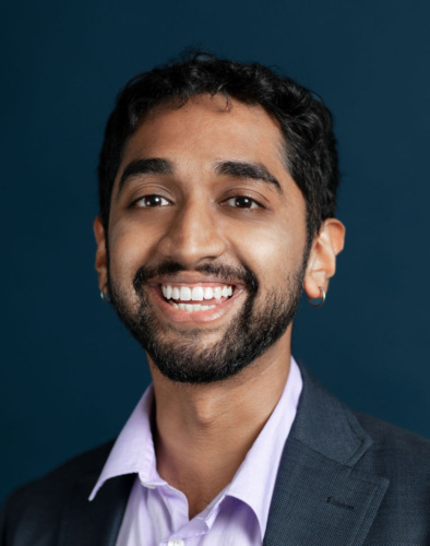 A man with short dark hair and a beard, wearing a gray suit jacket and a light purple shirt, smiles at the camera against a dark blue background.