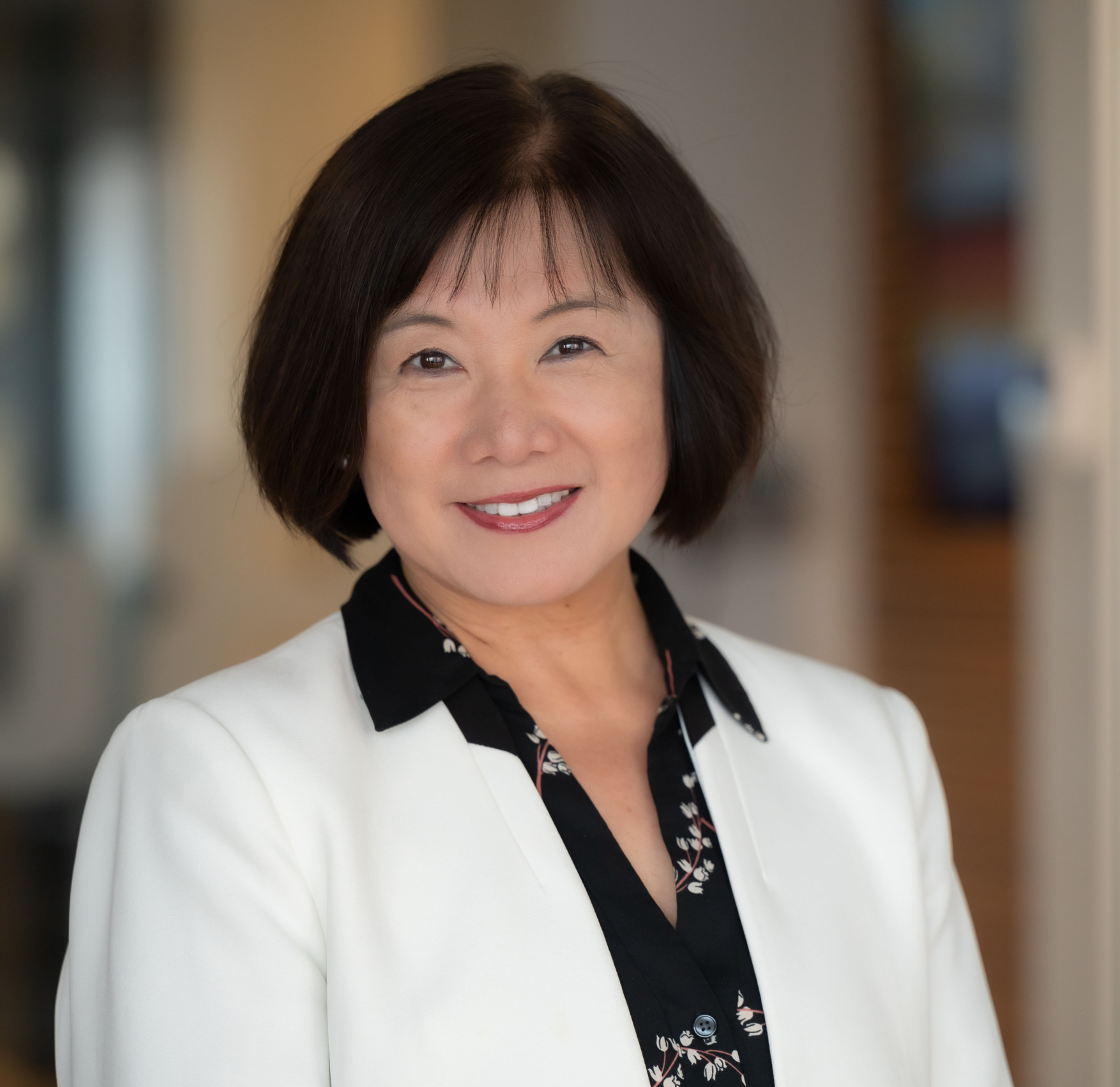 A woman with short dark hair wearing a white blazer and black blouse smiles at the camera in a professional indoor setting.