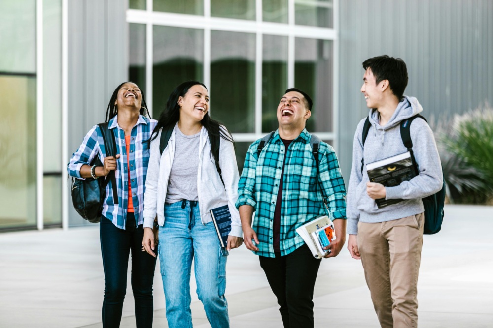 Four students walk together outside a modern building, smiling and laughing, carrying backpacks and books—excited to discuss opportunities like DACA Graduate School Loans for their academic futures.