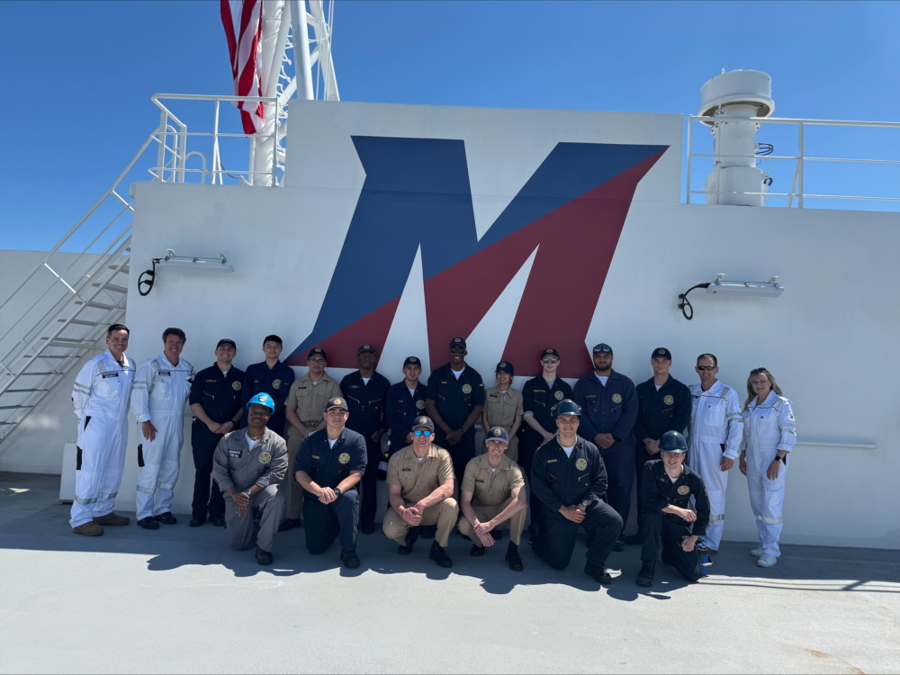 A group of uniformed crew members poses in front of a large “M” logo on the deck of a ship under a clear blue sky.
