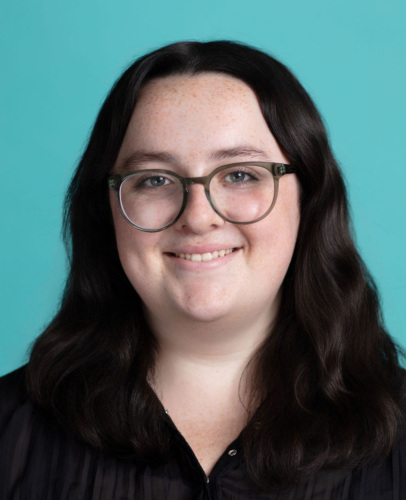 A person with long dark hair and glasses, wearing a black top, is smiling in front of a solid teal background.