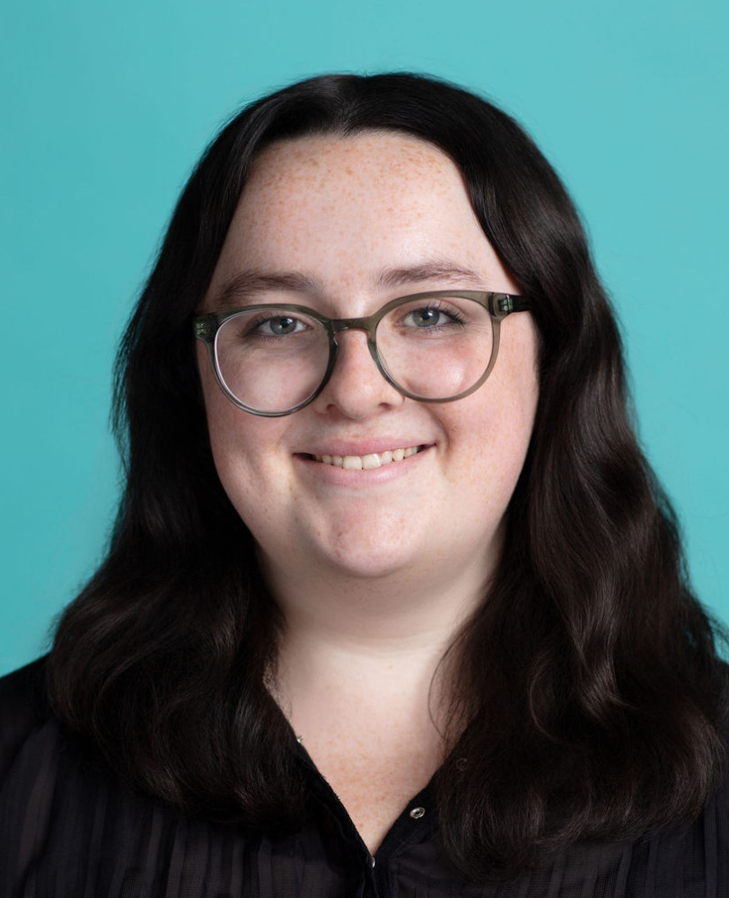 A person with long dark hair and glasses, wearing a black top, is smiling in front of a solid teal background.