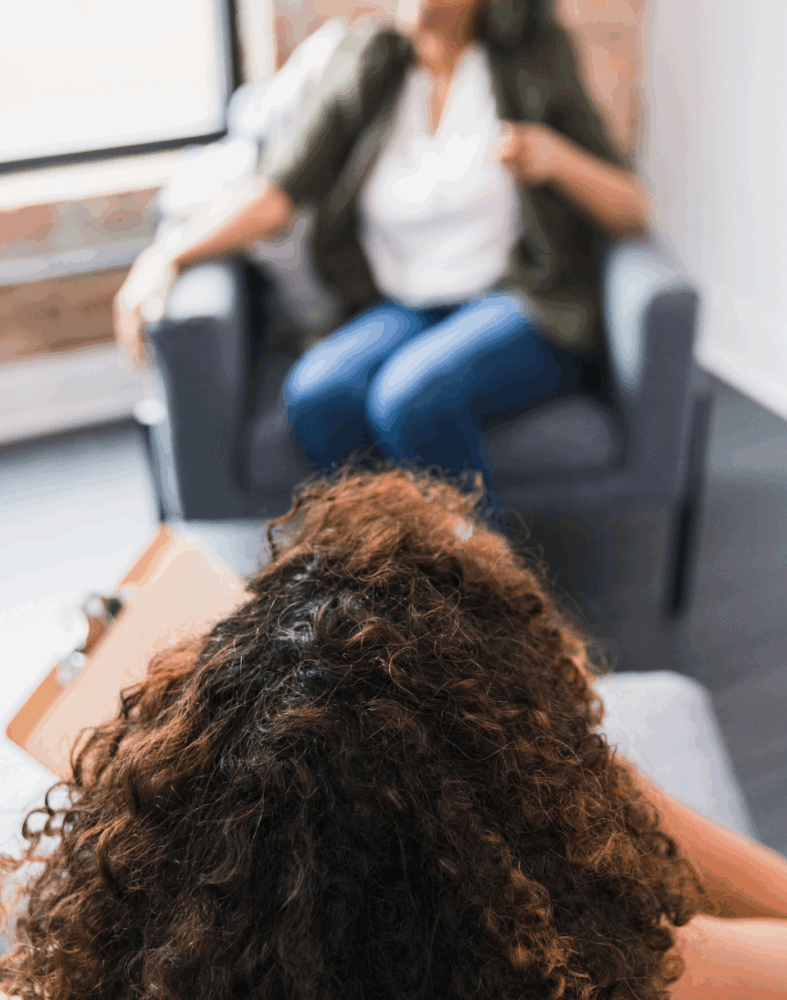 Two people sit across from each other in a brightly lit room, one taking notes on a clipboard, suggesting a counseling or therapy session.