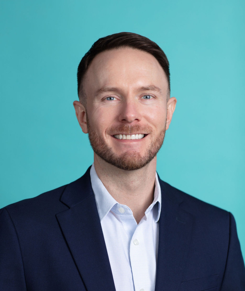 A man in a navy blazer and white shirt smiles at the camera against a plain turquoise background.