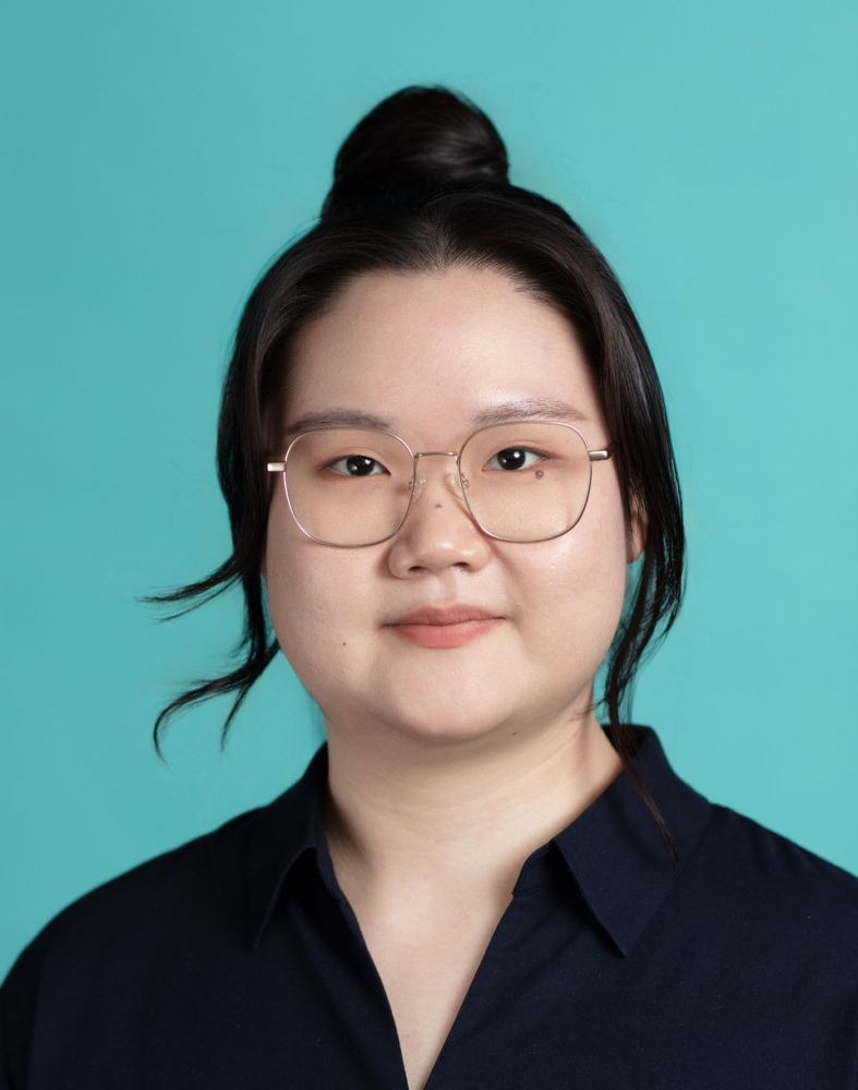 A person with long dark hair in a bun, wearing glasses and a navy blue collared shirt, poses in front of a solid turquoise background.
