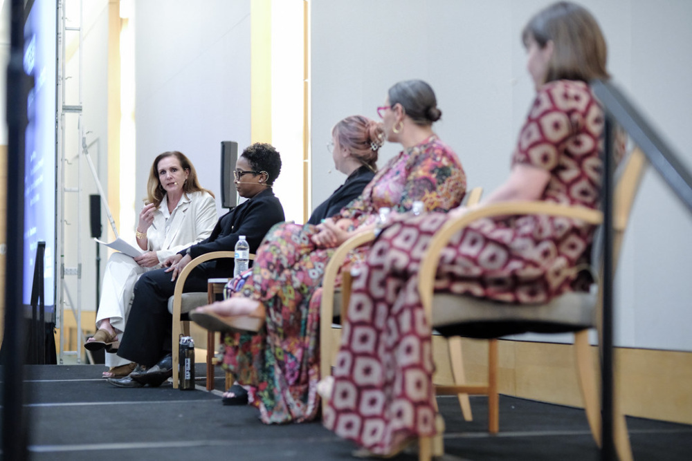 Five women sit on a stage engaged in a panel discussion, with one woman speaking about innovative funding models while the others listen intently.