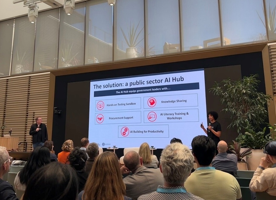 A presenter addresses an audience in a modern conference room, displaying a slide titled "The solution: a public sector AI hub" with five key focus areas.
